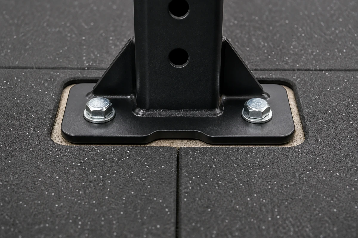 Tight close-up of a black power rack footplate bolted through a precise cutout in a 0.47-inch rubber tile into the concrete slab beneath, showing the wedge anchor bolt and quarter-inch clearance gap